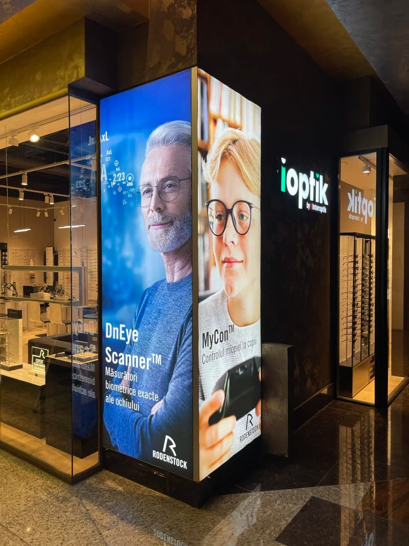 Casetă luminoasă cu față printată, amplasată în interiorul unui magazin optic. Prezintă imagini și text, fiind un element de semnalistică vizibil și atractiv.