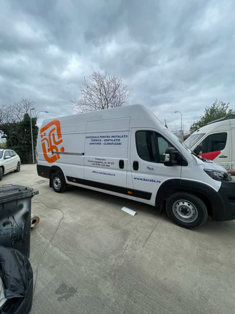 O autoutilitară albă cu colantare auto din vinil, expunând text și grafică colorată. Vehiculul este parcat într-un mediu exterior.